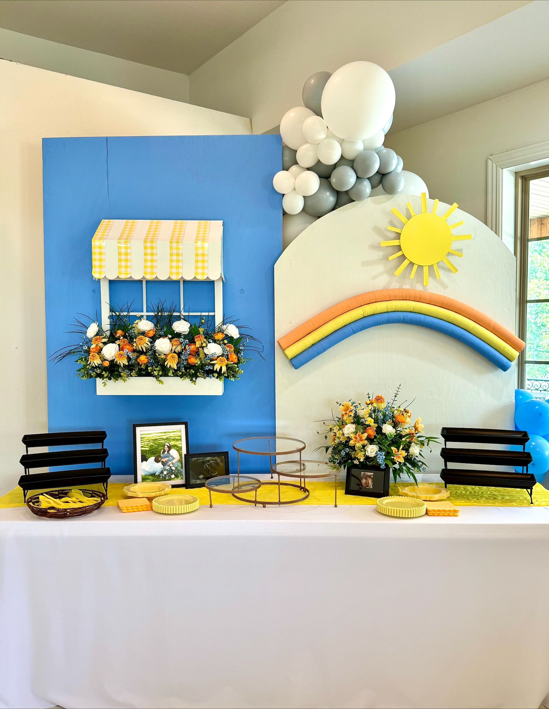 Balloon-decorated dessert table with blue and rainbow backdrops, flowers, and gold trays
