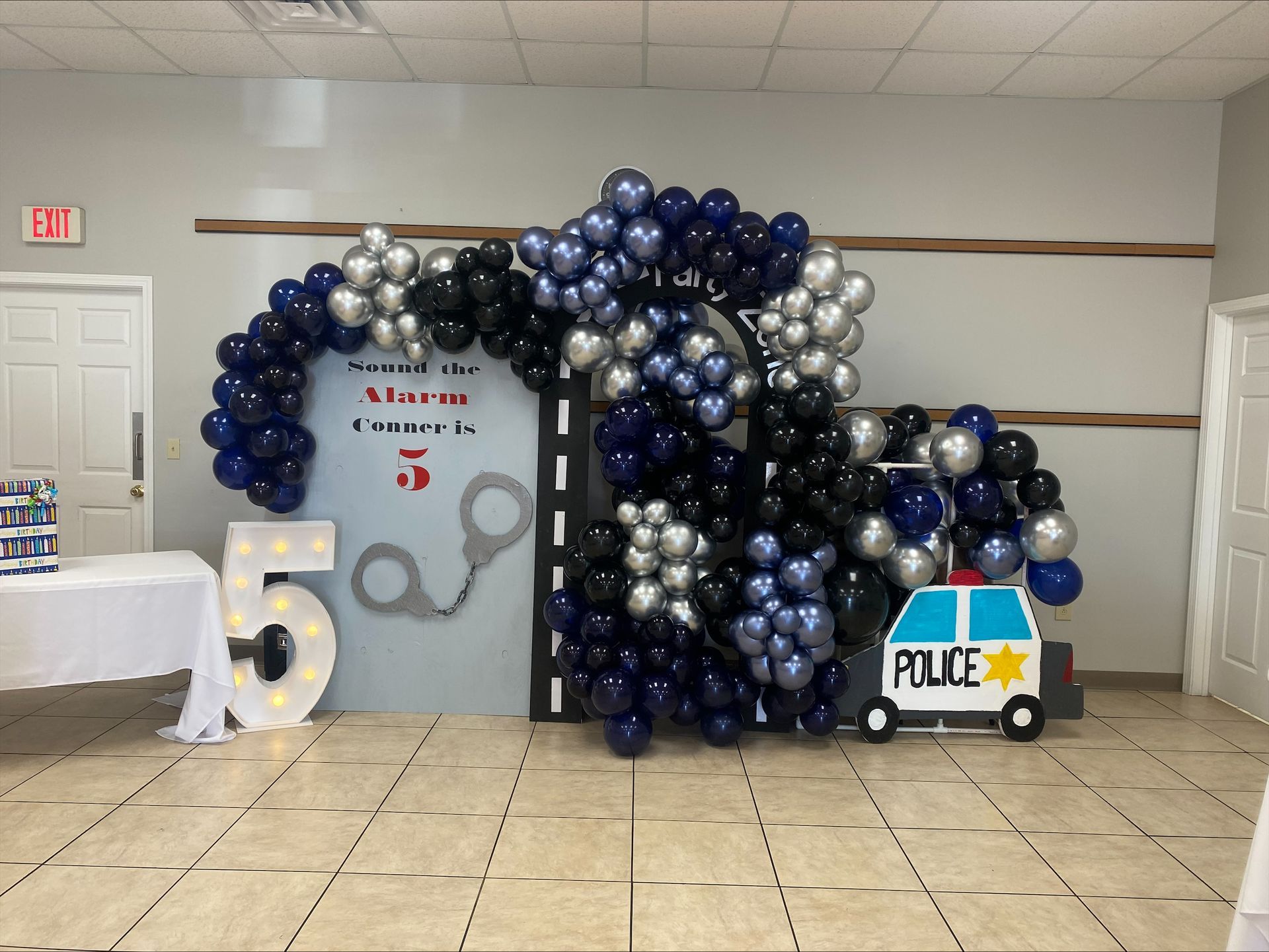 Balloon arch over a dance-themed display with a 15 sign and a small police car prop in a hallway