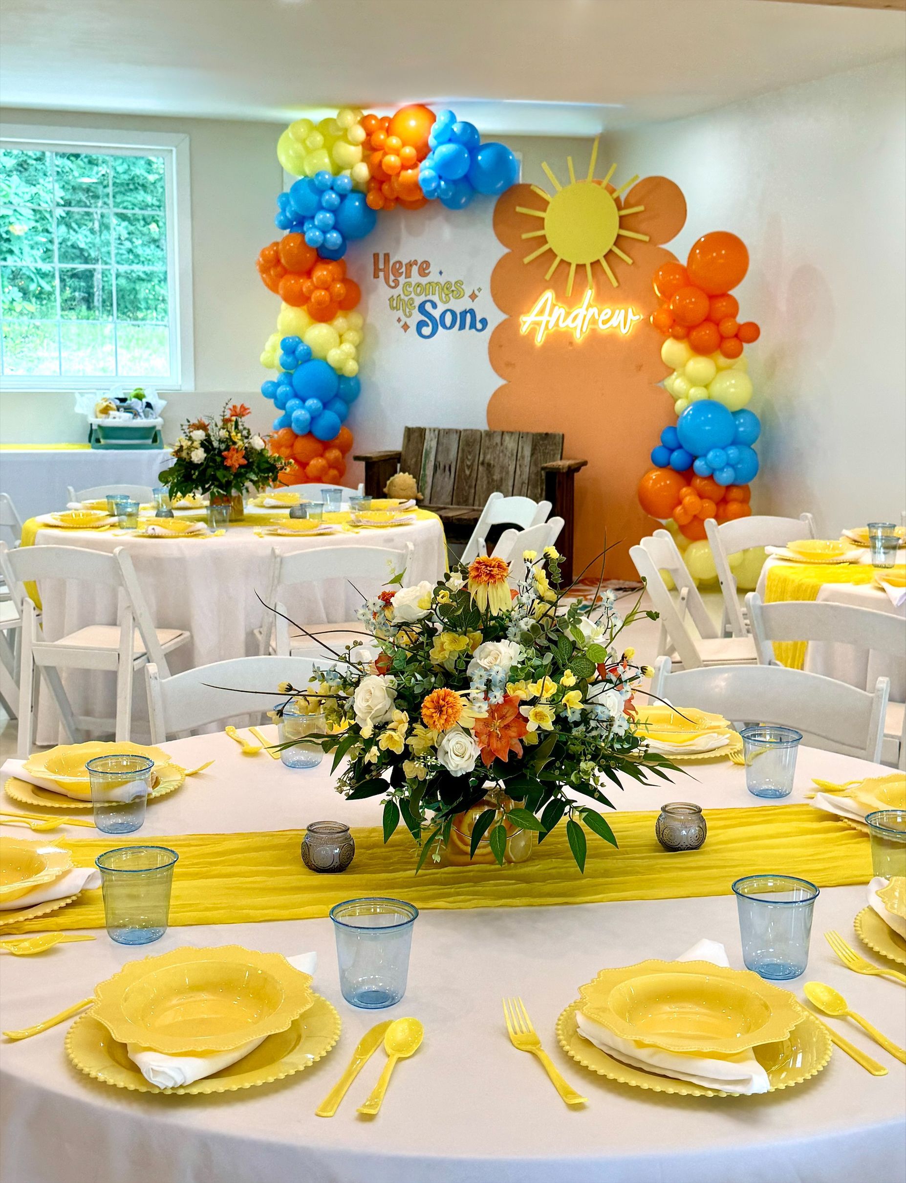 Brightly decorated banquet table with yellow napkins and blue-orange balloon arch backdrop at an indoor celebration