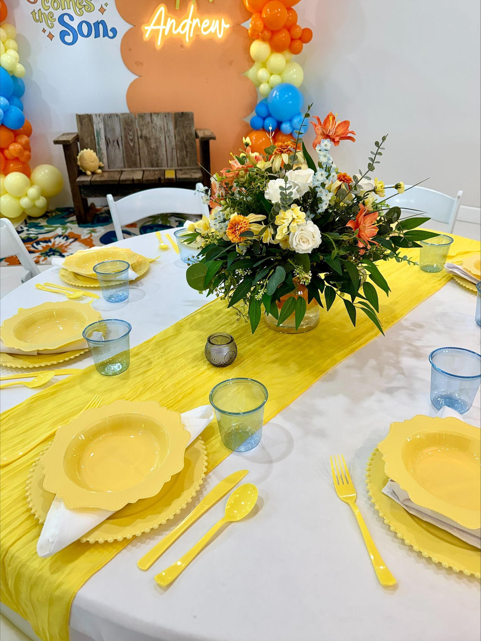 Yellow-themed birthday table with floral centerpiece, plates, cups, and balloons in the background
