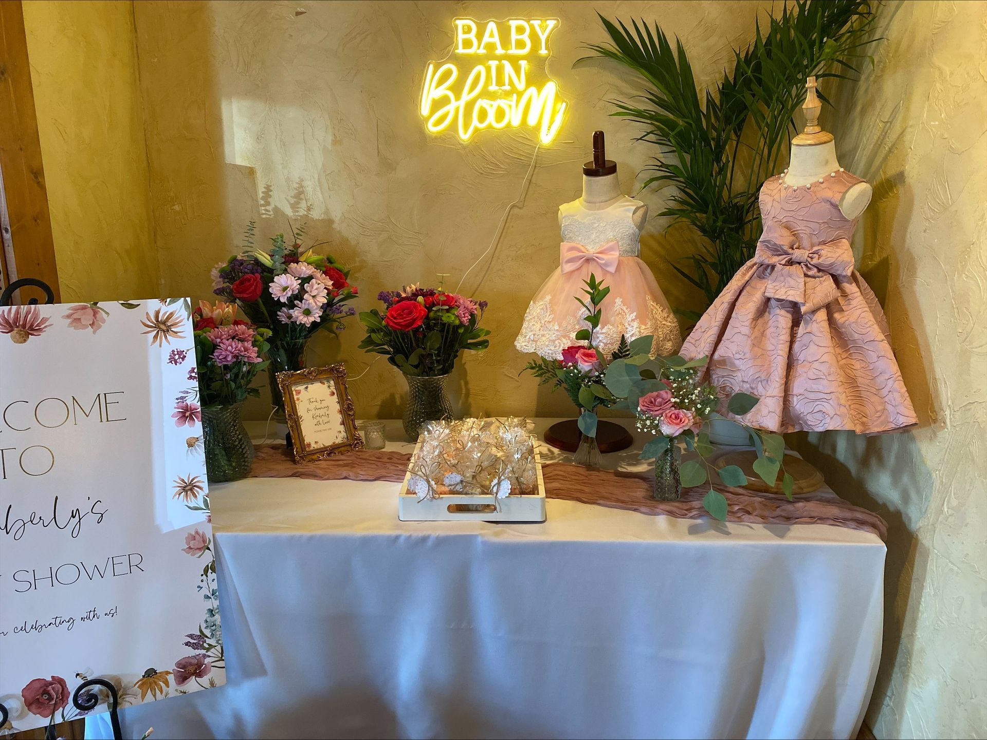 Baby shower dessert table with cake, flowers, gifts, and a pink dress display under a neon sign