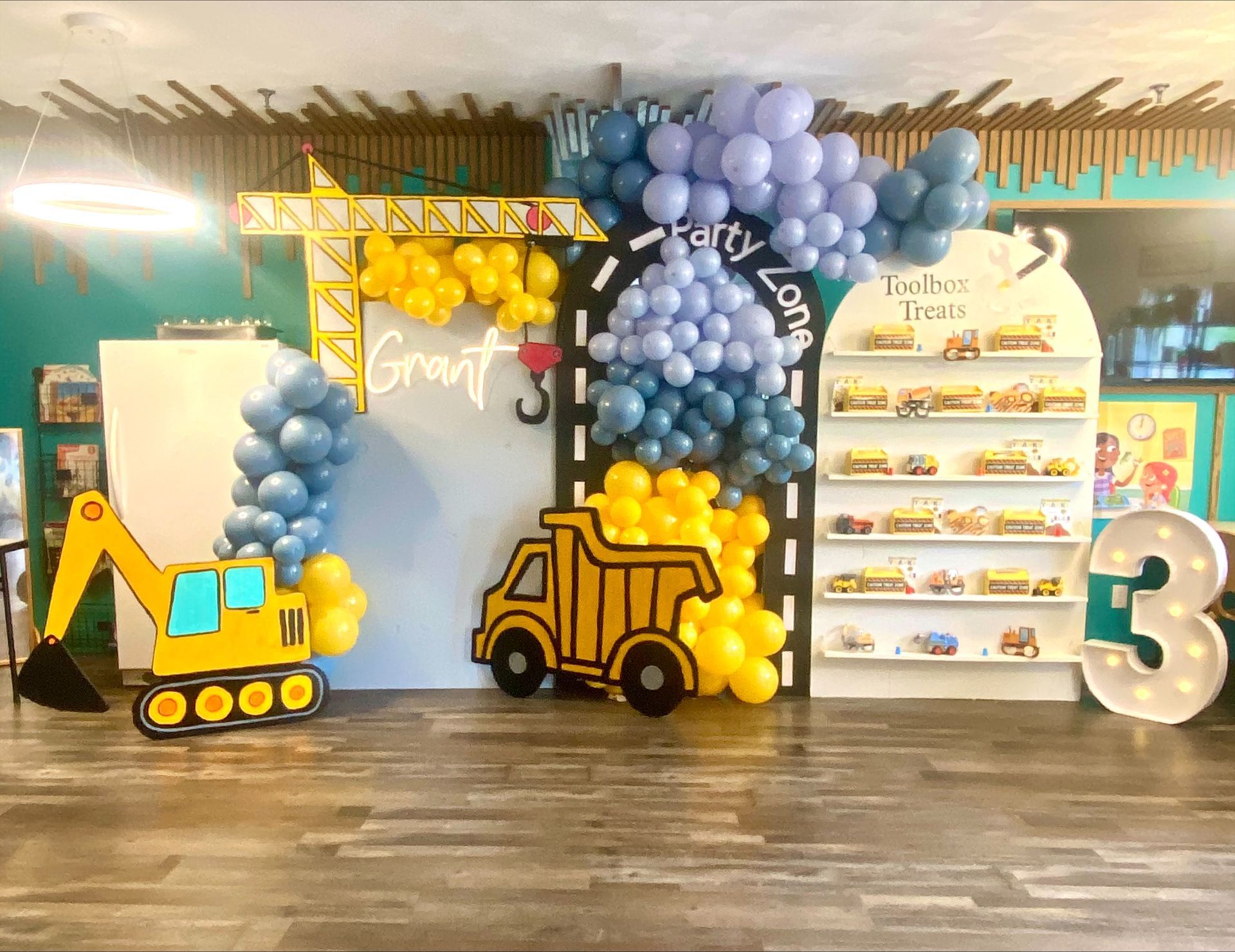 Construction-themed birthday display with yellow, blue, and lavender balloons, toy trucks, and a large number 3 backdrop