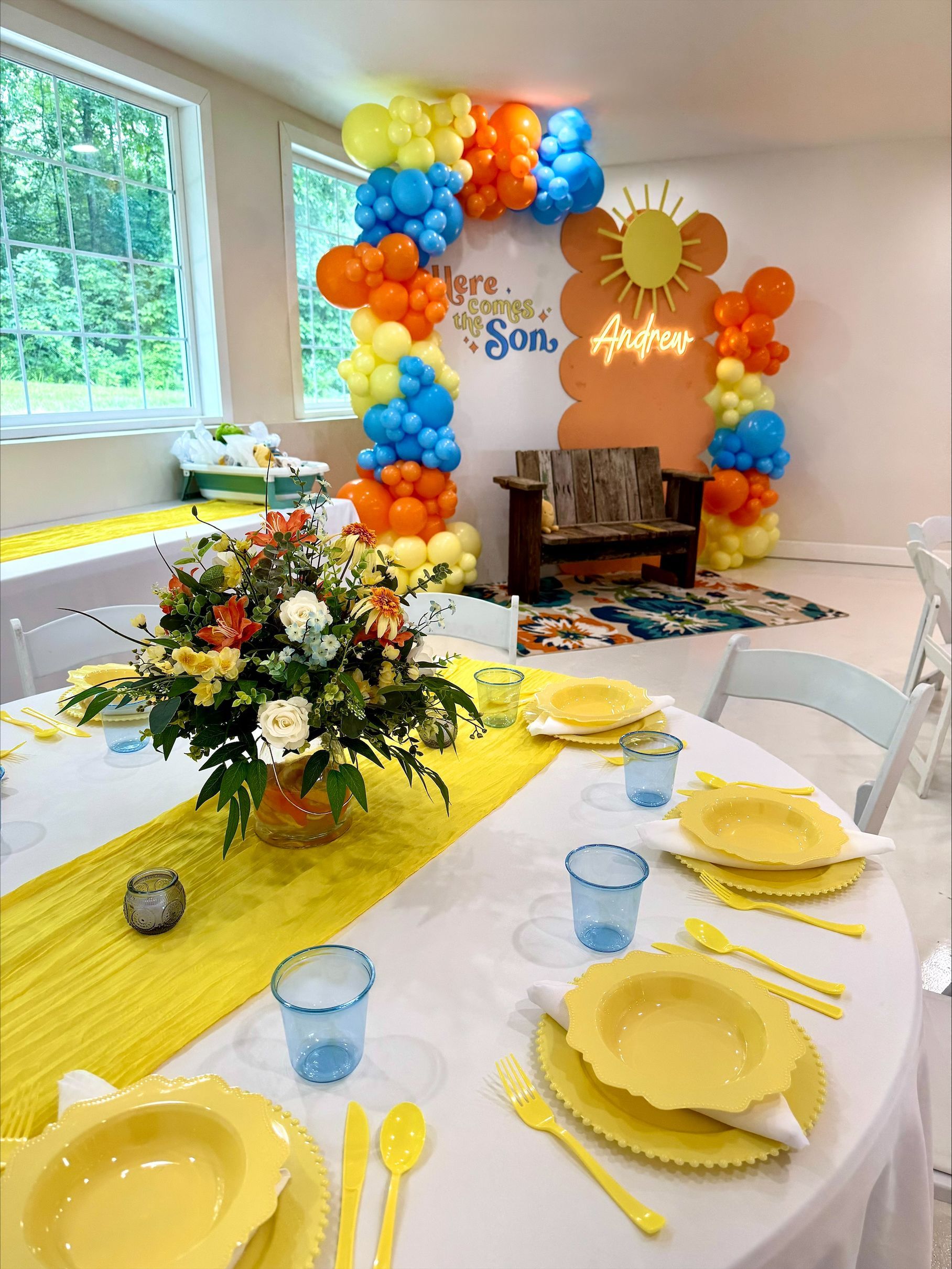 Decorated party table with yellow dishes, blue cups, orange-blue balloons, and a floral centerpiece by a fireplace