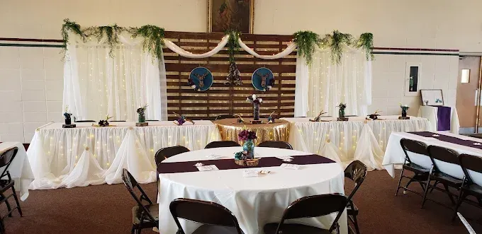 Decorated banquet hall with round tables, white chair covers, and a stage backdrop with draped greenery