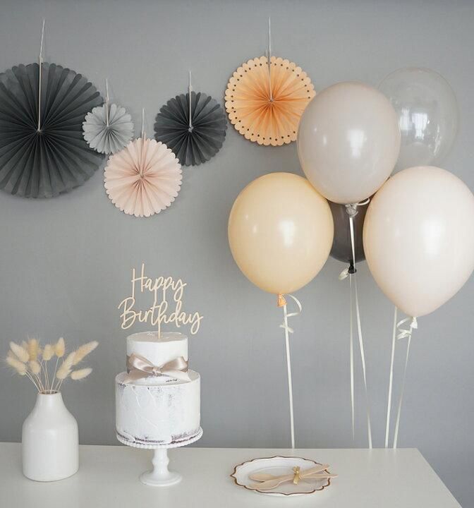 Birthday dessert table with white cake, gold balloons, and gray paper fan decorations against a gray wall