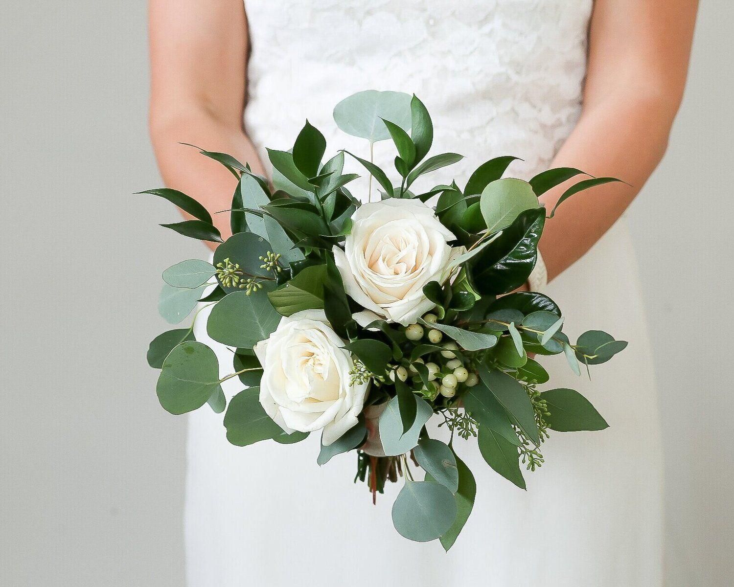 Bridal bouquet of white roses and greenery held by a person in a white dress