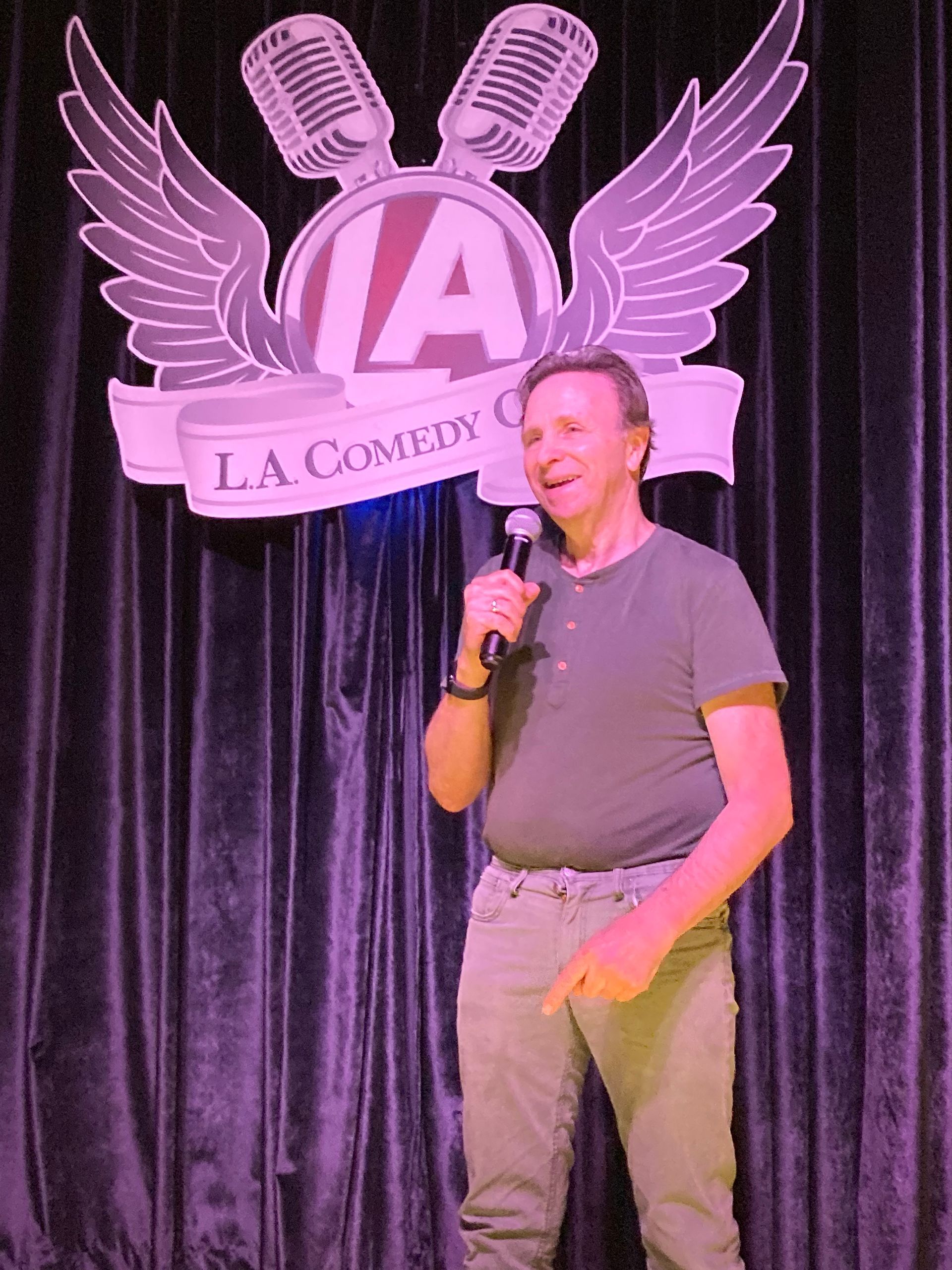 Man speaking into a microphone on a lit stage with purple curtains and an LA Comedy Club sign behind him