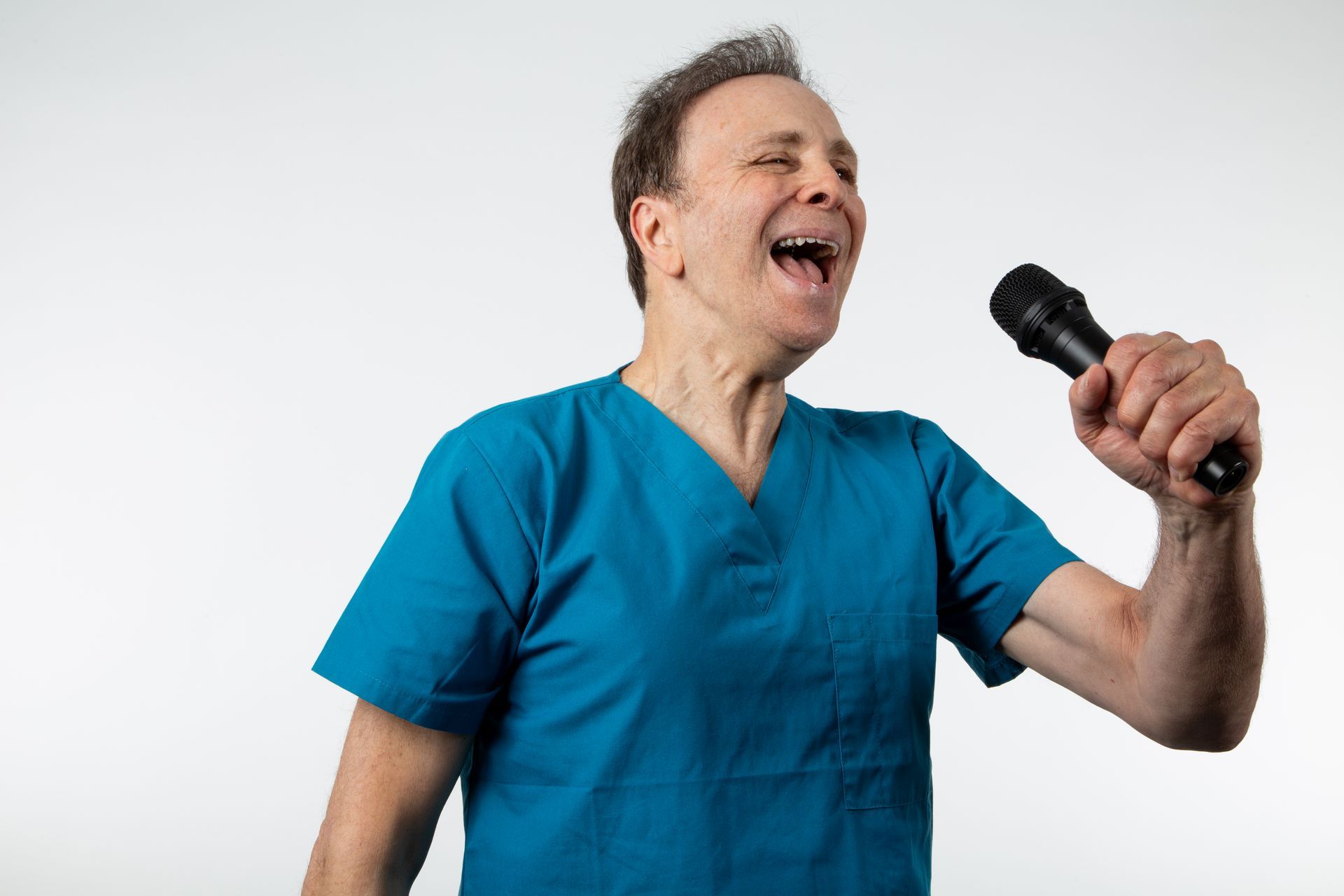 Man in blue scrubs singing into a microphone, mouth open, against a white background