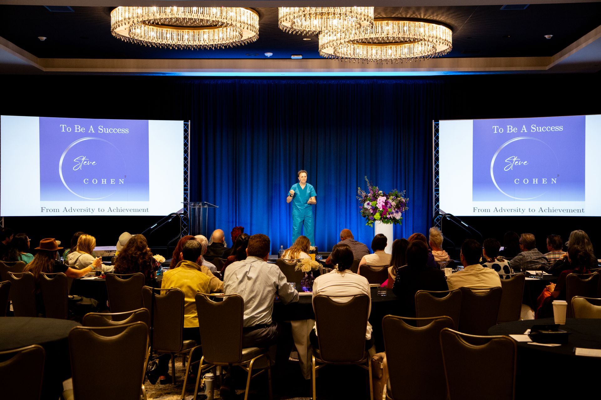 Speaker on stage at conference, addressing audience in a ballroom with blue curtains and large screens.