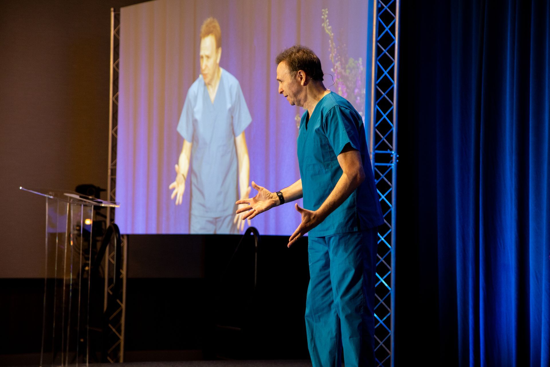 Speaker on stage in blue scrubs, gesturing beside a projected image during a presentation