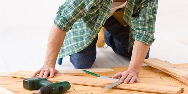A man is kneeling down on the floor cutting a piece of wood.
