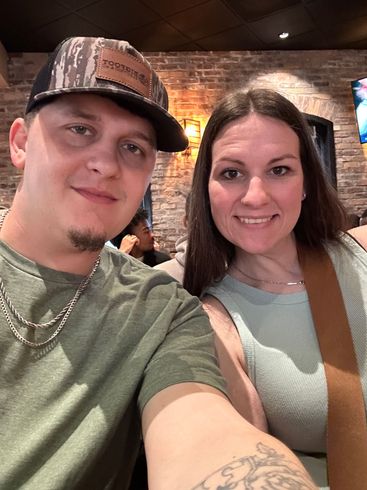 Two smiling people taking a selfie indoors under warm lighting, wearing casual clothes and caps.