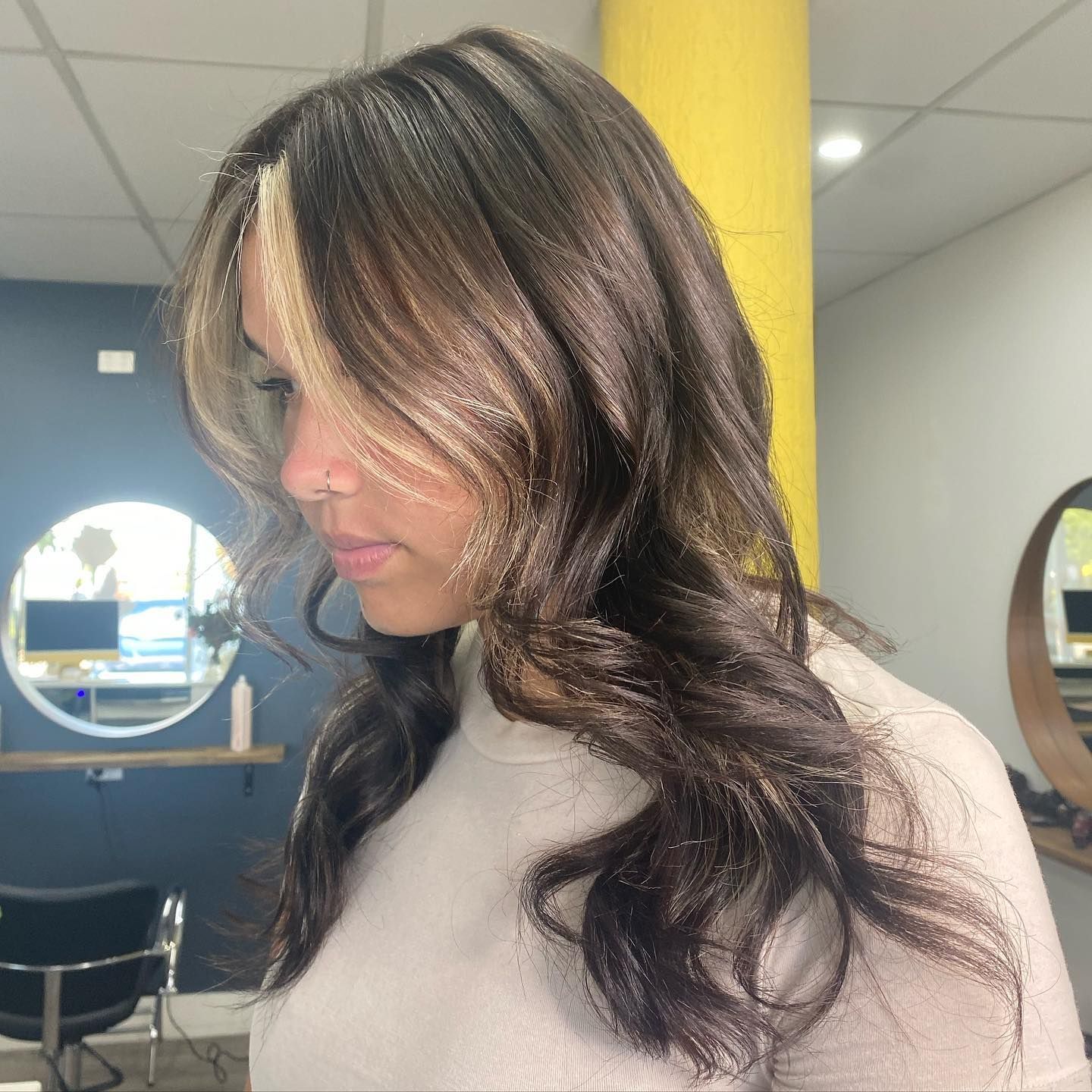 Woman With Long Wavy Dark Hair In A Light Gray Top, Shown In Profile Indoors Near Yellow And Blue Walls — Oh So Blonde Hair Studio in Kawana, QLD