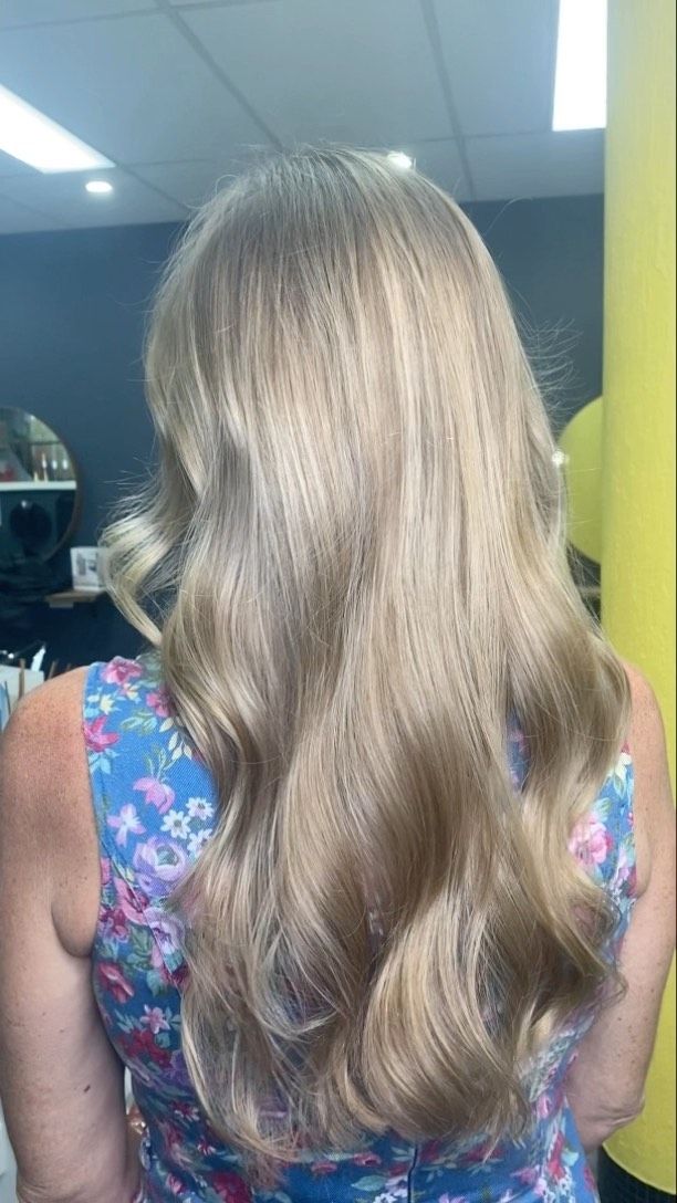 Person Seen From Behind With Long Wavy Blonde Hair, Wearing A Floral Dress In A Dim Indoor Room — Oh So Blonde Hair Studio in Kawana, QLD