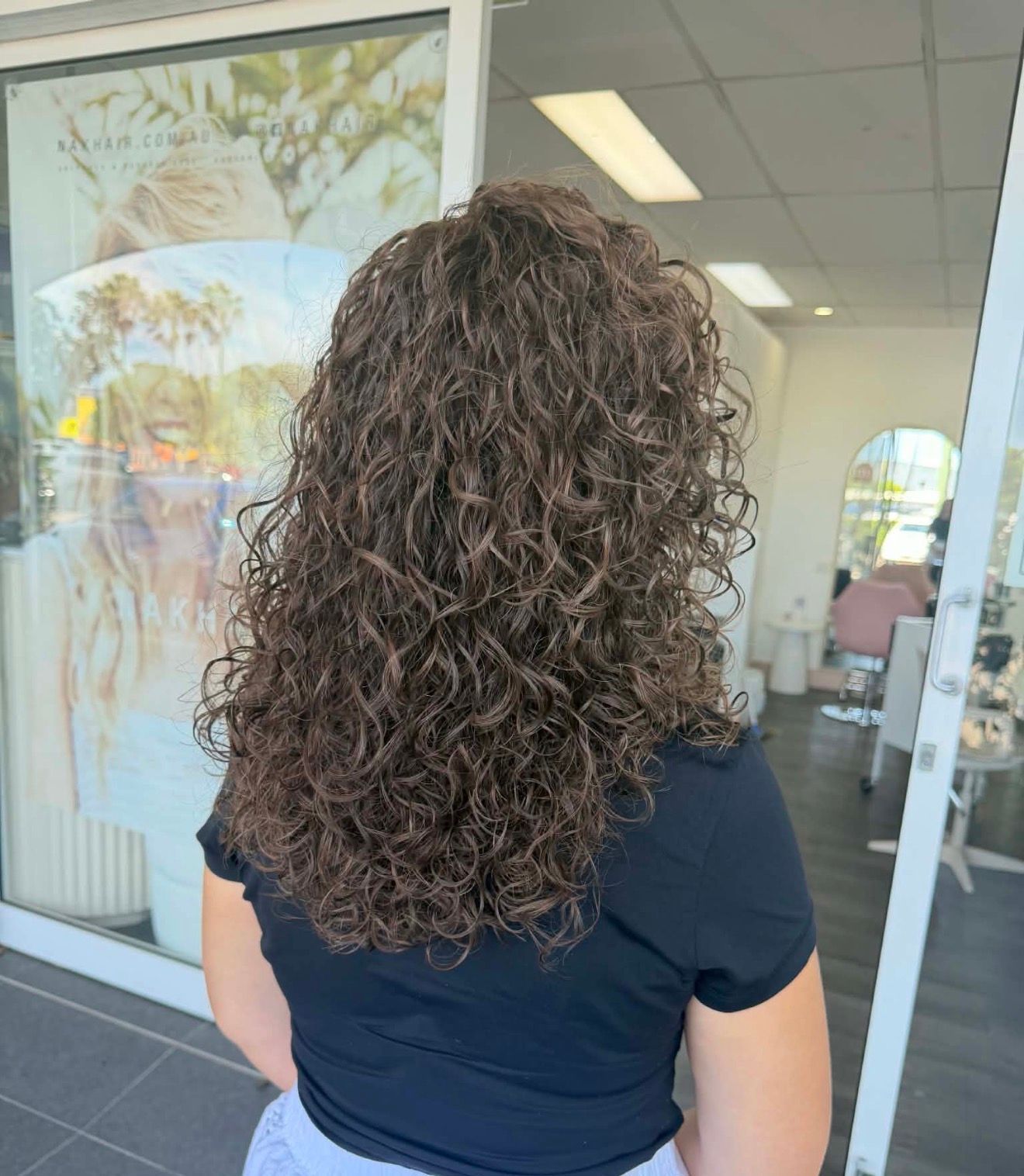 Back View Of A Person With Curly Dark Hair In A Black Salon Cape Indoors — Oh So Blonde Hair Studio in Kawana, QLD