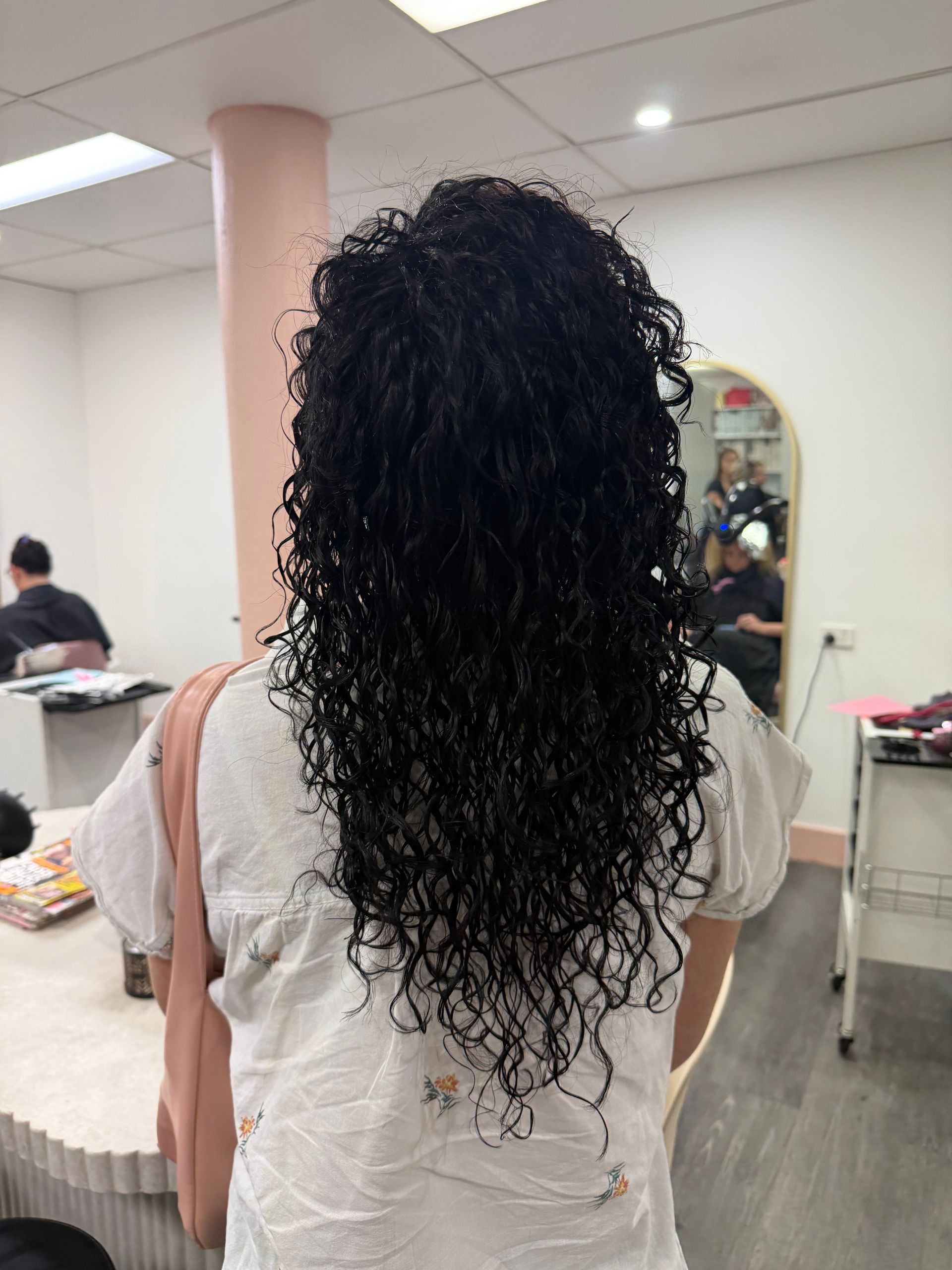 Person With Long Curly Dark Hair Seen From Behind In A Salon, Wearing A Light Top With A Shoulder Bag — Oh So Blonde Hair Studio in Kawana, QLD