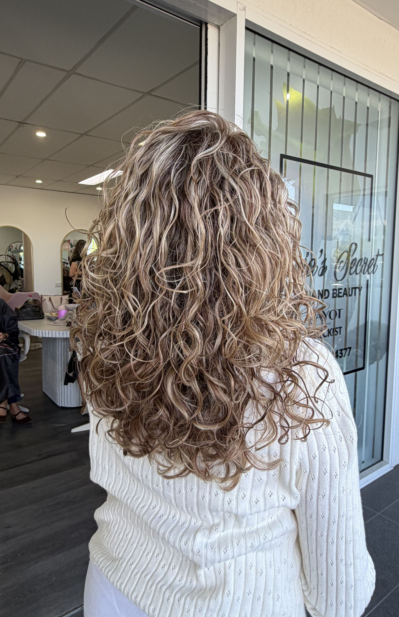 Woman With Curly Highlighted Hair Seen From Behind In A Salon Lobby, Wearing A White Sweater — Oh So Blonde Hair Studio in Kawana, QLD