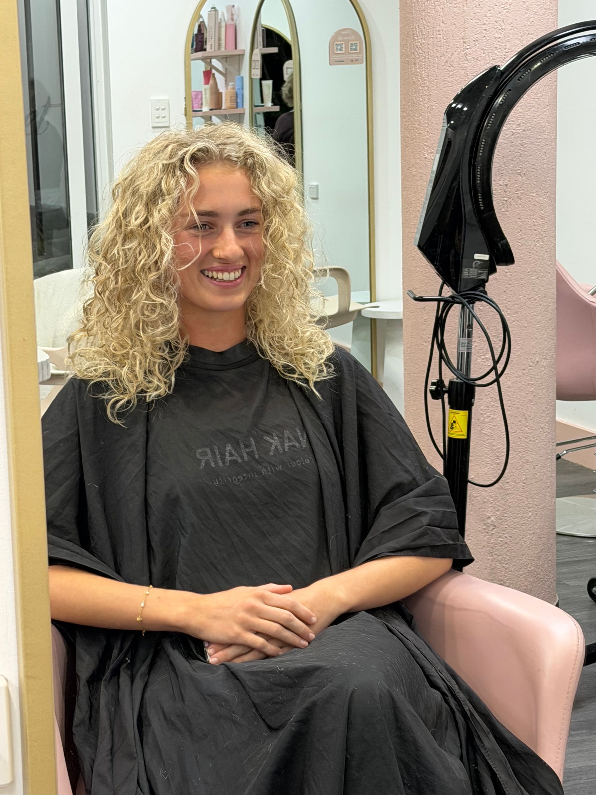 Person With Curly Blonde Hair Smiling In A Salon Chair, Wearing A Black Cape With A Mirror Behind Them — Oh So Blonde Hair Studio in Kawana, QLD