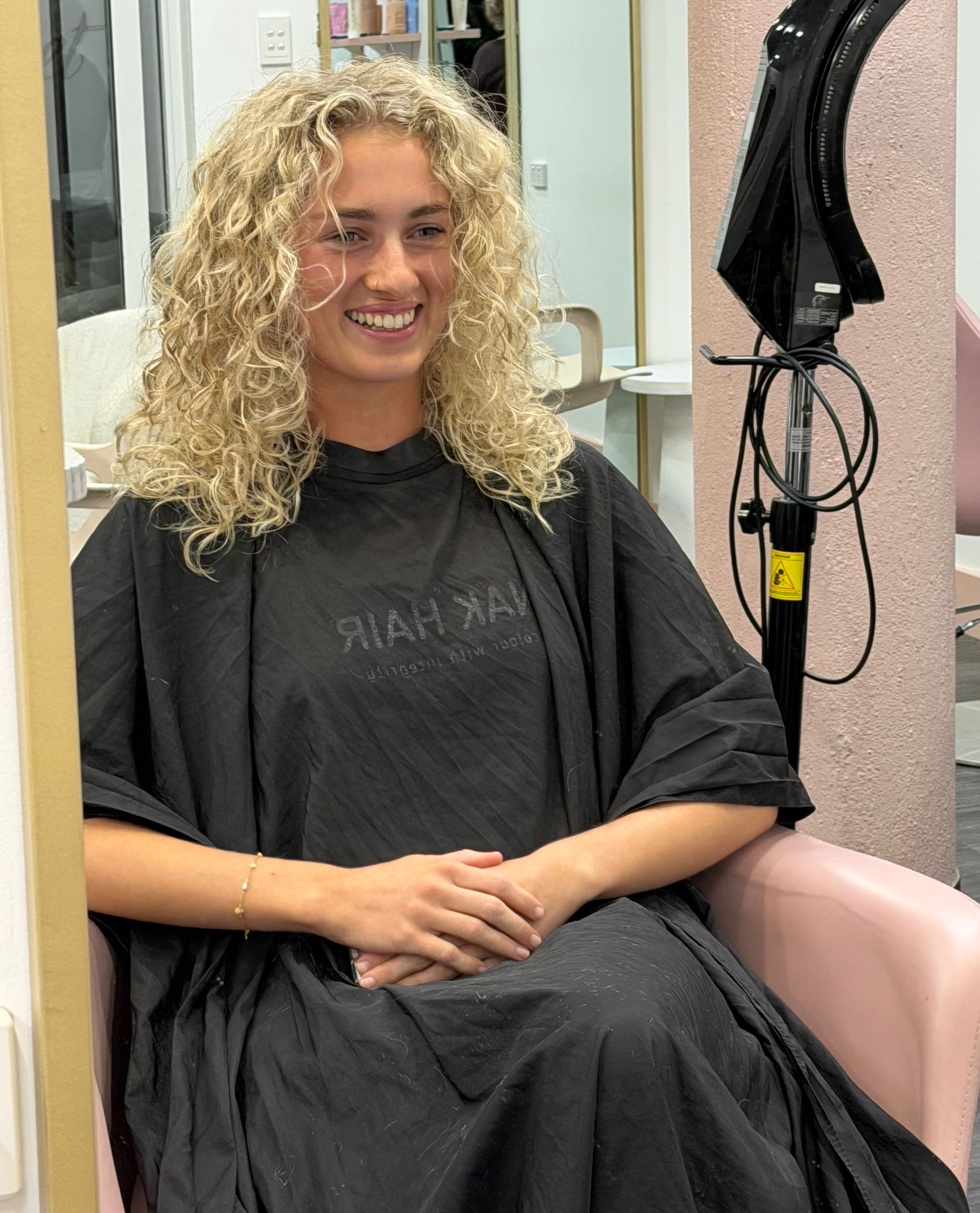 Woman With Curly Blonde Hair Smiling In A Salon Chair, Wearing A Black Cape And Dark Shirt — Oh So Blonde Hair Studio in Kawana, QLD