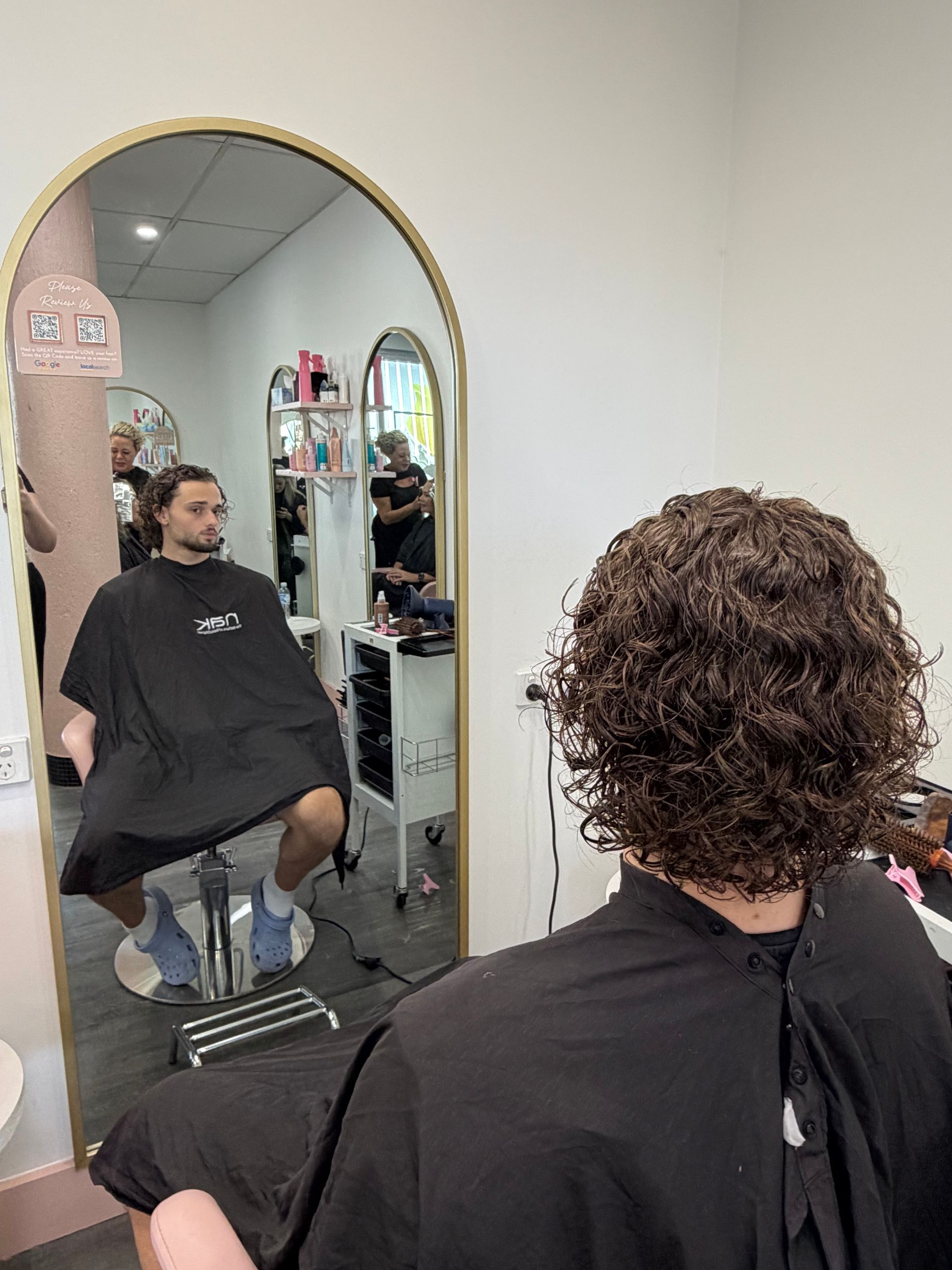 Person Seated In A Salon Chair, Seen In A Mirror During A Haircut, With Curly Hair And A Black Cape — Oh So Blonde Hair Studio in Kawana, QLD