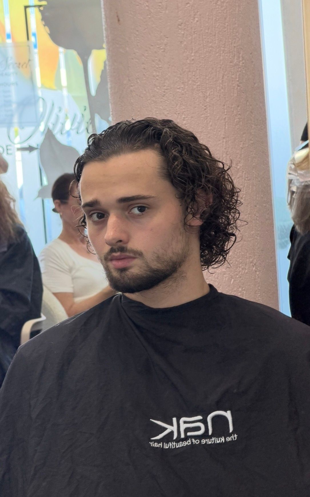 Person With Curly Dark Hair And Beard, Wearing A Black T-shirt, Seated Indoors Against A Colorful Wall — Oh So Blonde Hair Studio in Kawana, QLD
