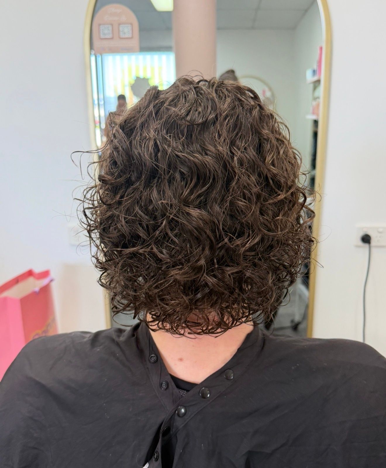 Back View Of A Person With Curly Brown Hair In A Black Cape Inside A Salon — Oh So Blonde Hair Studio in Kawana, QLD