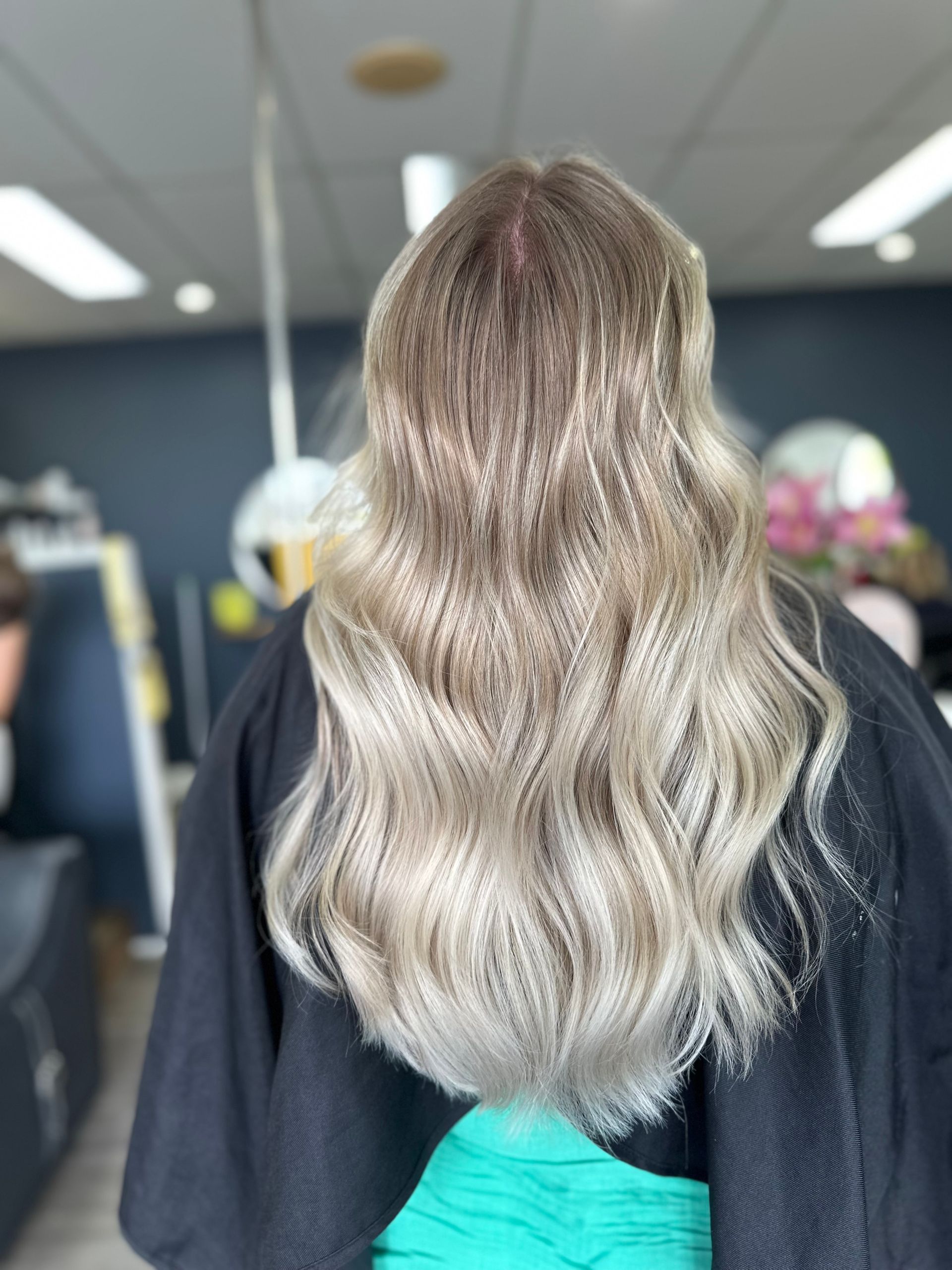 Long Wavy Blonde Hair Viewed From Behind In A Salon Chair With A Black Cape And Turquoise Clothing Visible — Oh So Blonde Hair Studio in Kawana, QLD