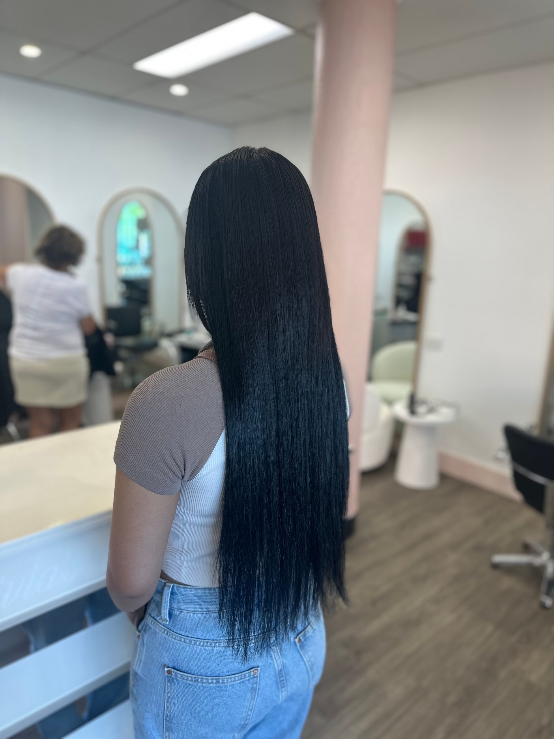 Person With Long Black Hair Standing In A Salon, Facing A Mirror Near A Pastel Pink Column — Oh So Blonde Hair Studio in Kawana, QLD
