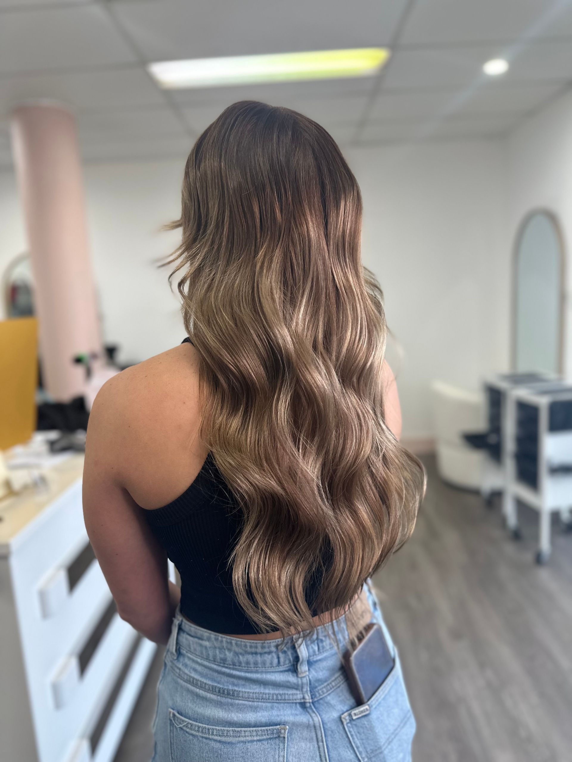 Woman With Long Wavy Hair Standing In A Salon, Seen From Behind In A Black Top And Light Jeans — Oh So Blonde Hair Studio in Kawana, QLD