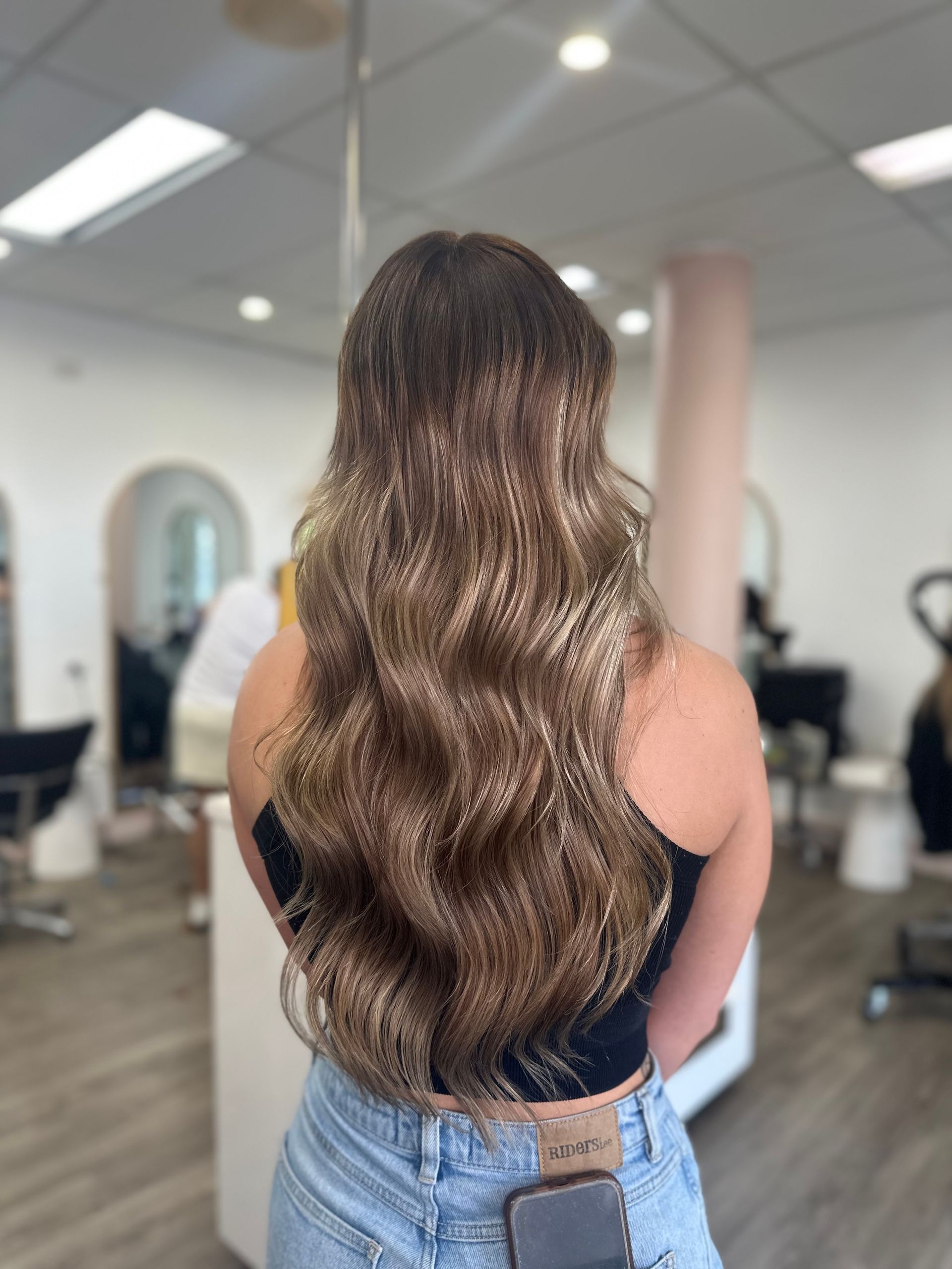 Woman With Long Wavy Brown Hair Standing In A Salon, Seen From Behind, Wearing A Black Top And Jeans — Oh So Blonde Hair Studio in Kawana, QLD