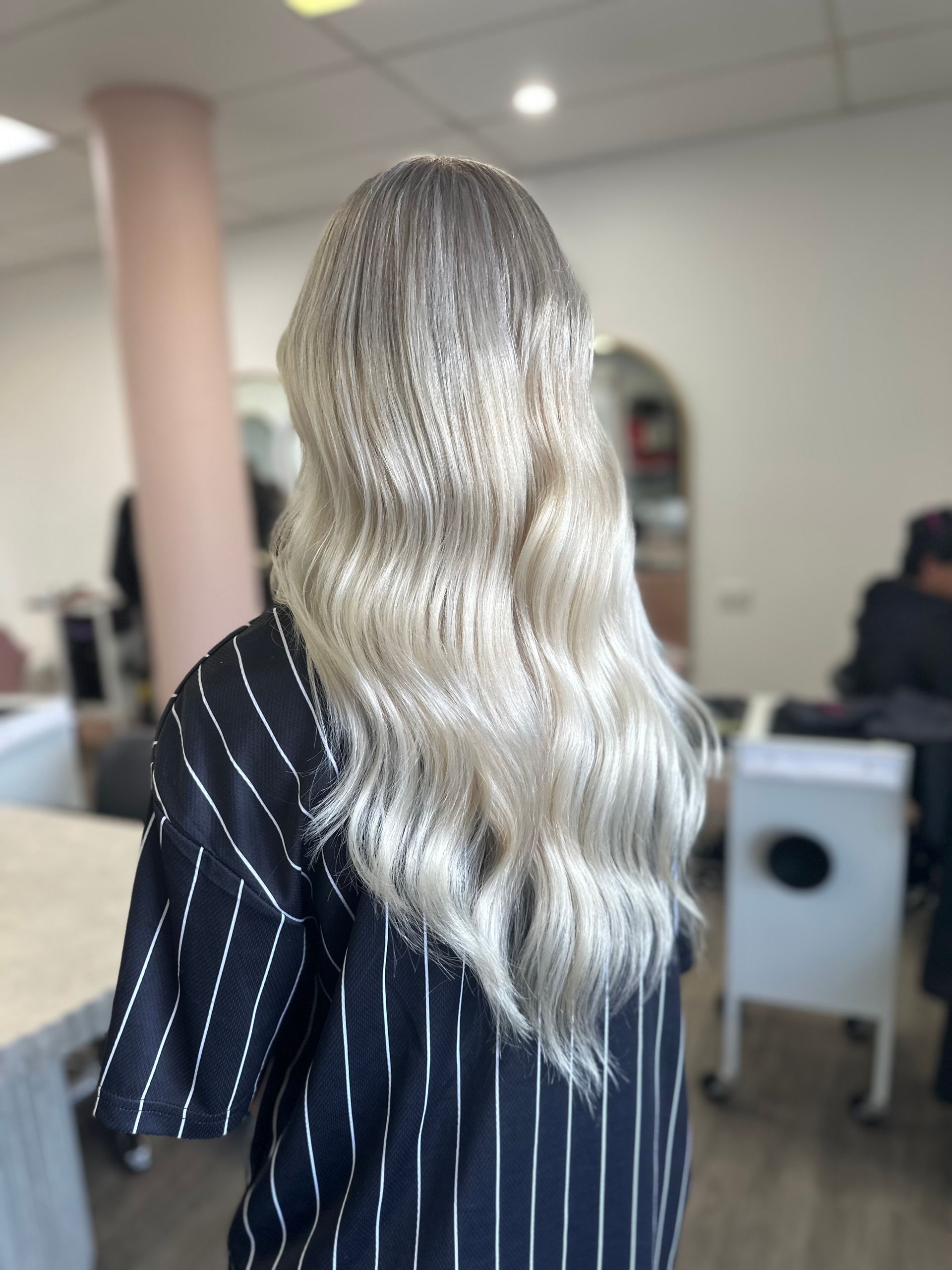 Person With Long Wavy Blonde Hair, Seen From Behind In A Striped Black Shirt Indoors — Oh So Blonde Hair Studio in Kawana, QLD