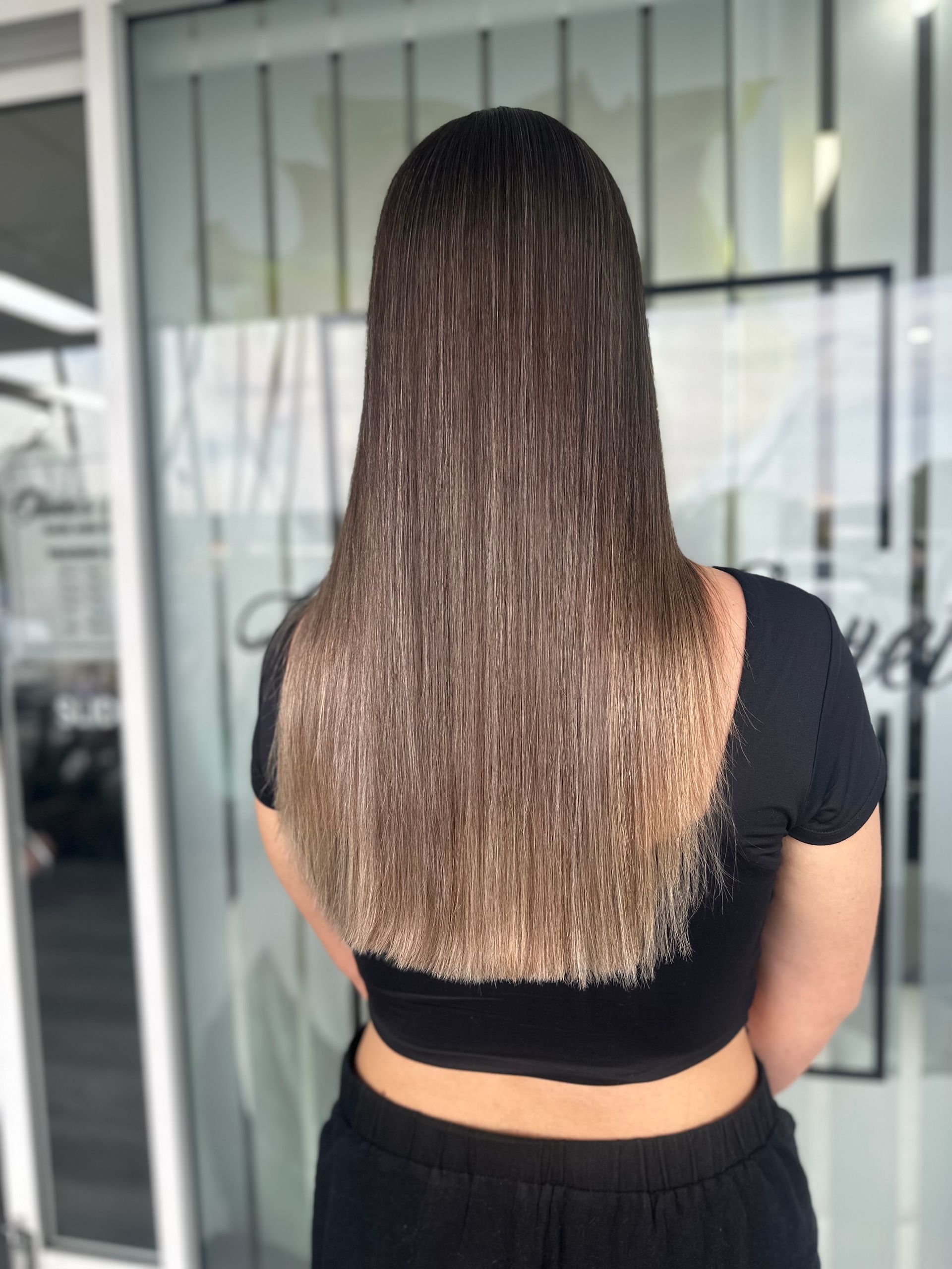 Person Seen From Behind With Long Straight Hair, Standing Indoors In A Black Top And Pants — Oh So Blonde Hair Studio in Kawana, QLD