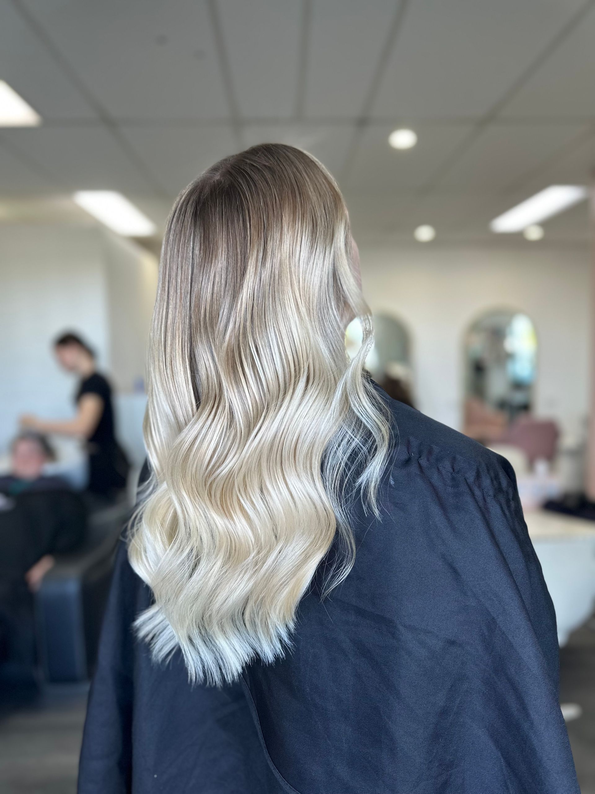 Blonde Wavy Hair Viewed From Behind In A Salon, Wearing A Dark Jacket With Mirrors And People In The Background — Oh So Blonde Hair Studio in Kawana, QLD
