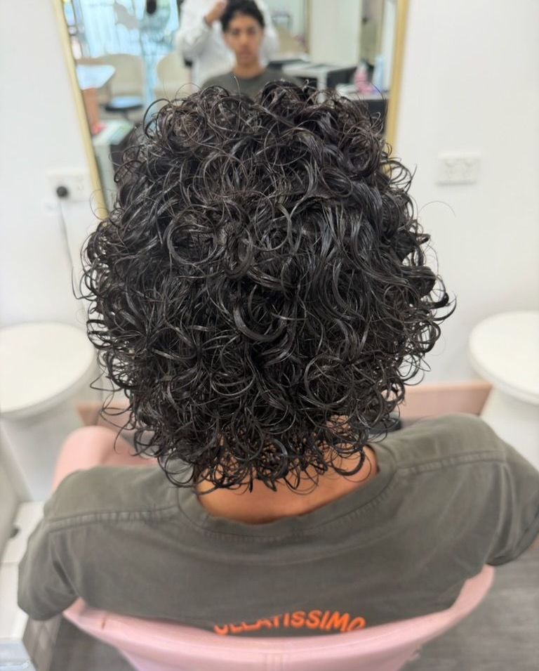 Back View Of A Person With Short Curly Dark Hair In A Salon Chair, Facing A Mirror — Oh So Blonde Hair Studio in Buddina, QLD