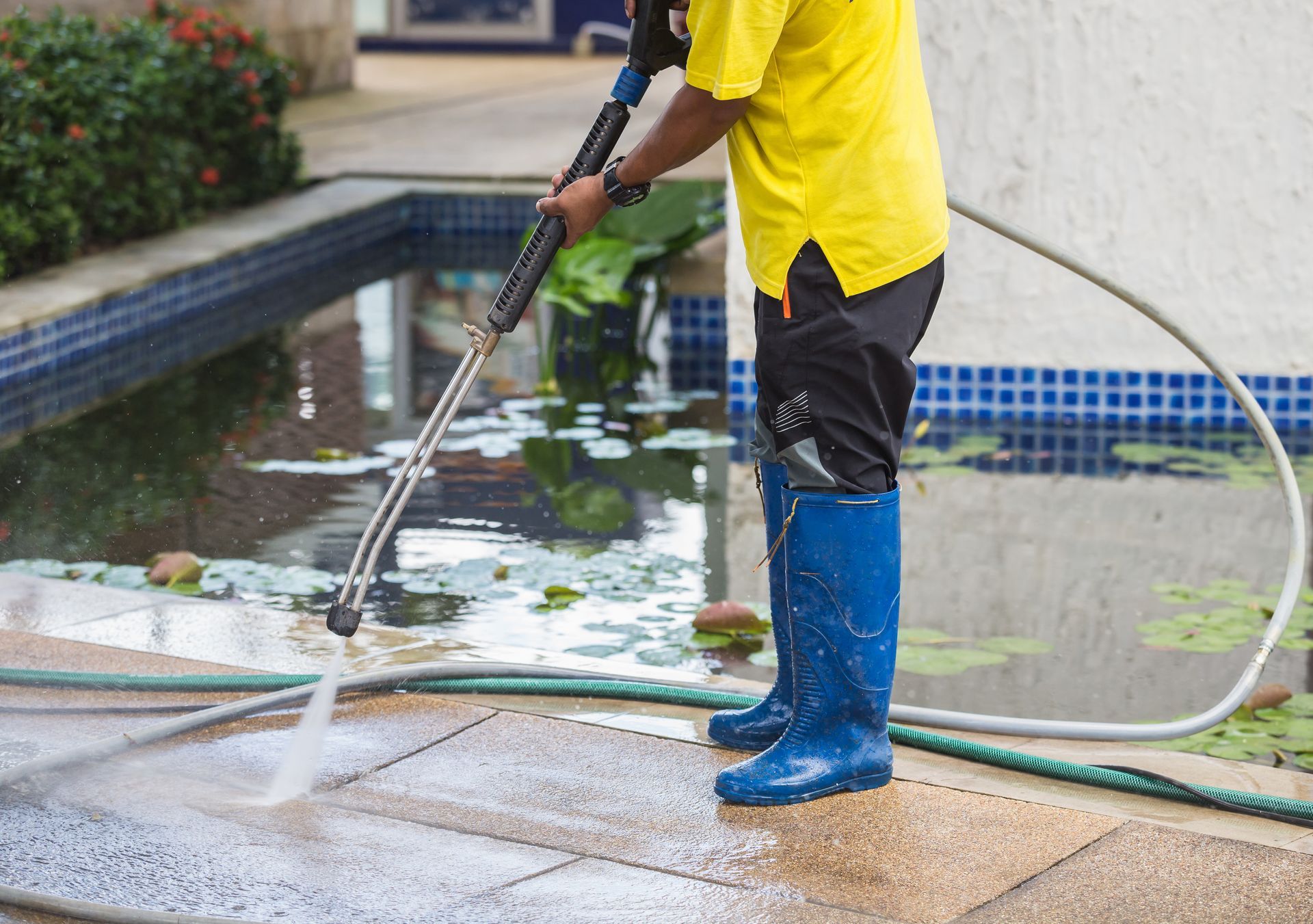 A person in a yellow shirt is pressure-washing a poolside patio.