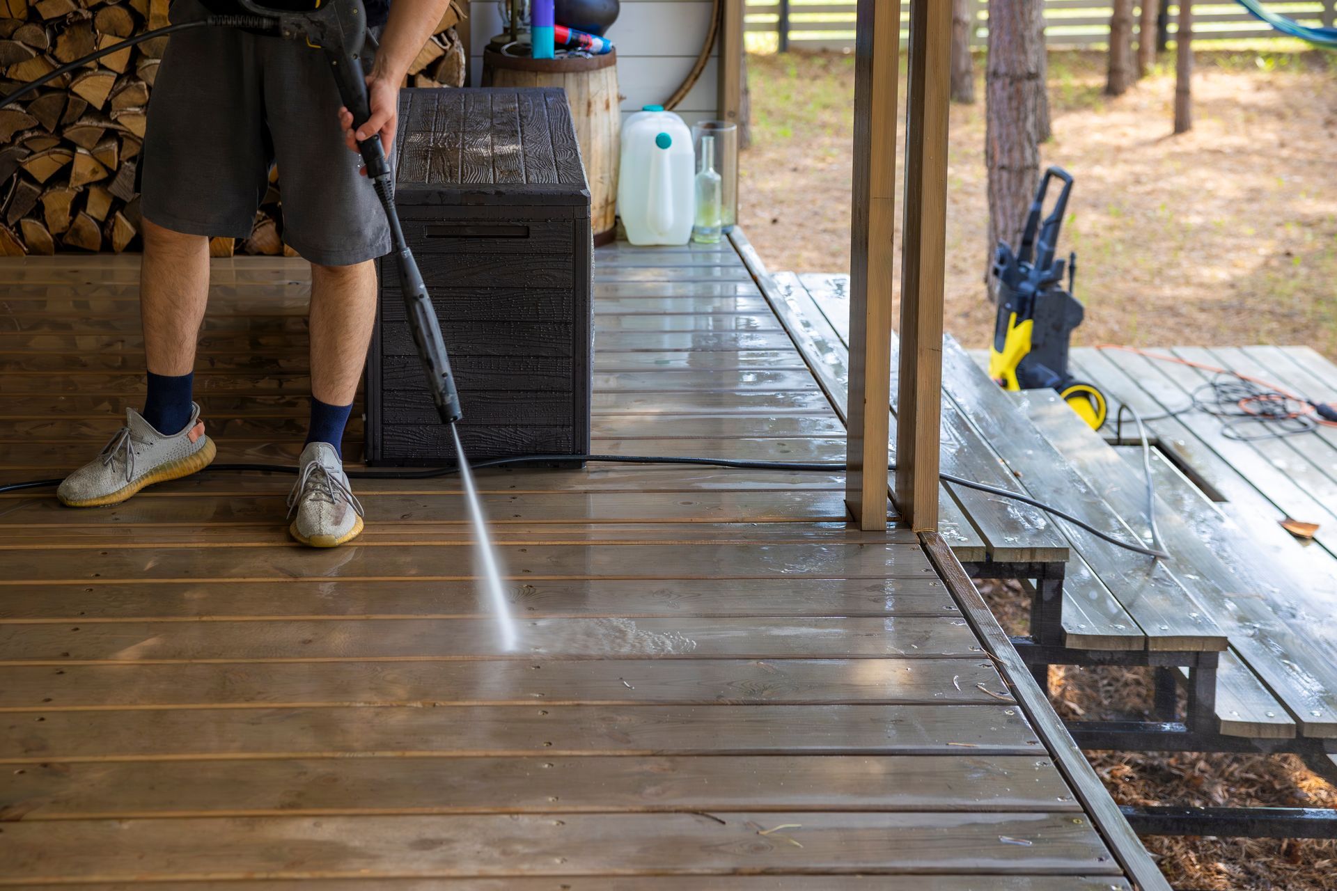 Person power-washing a wooden deck beside a black cabinet and yellow tools in an outdoor work area.
