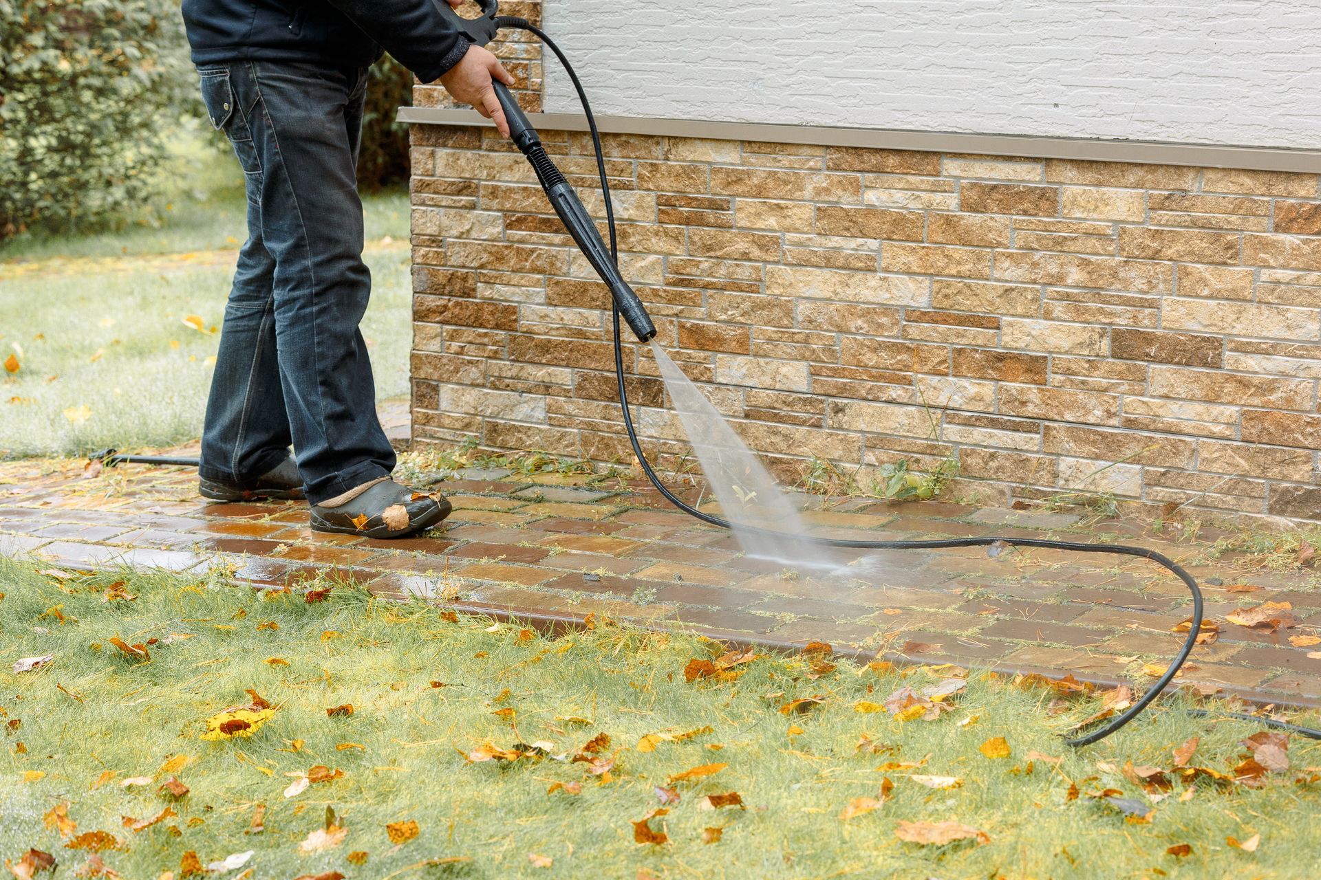 Person pressure-washing a stone wall and wet sidewalk outdoors in fall.