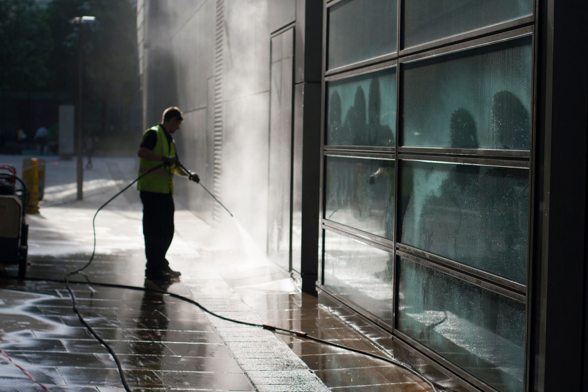 Worker pressure-washing a glass building exterior in bright sunlight, with mist and wet pavement.