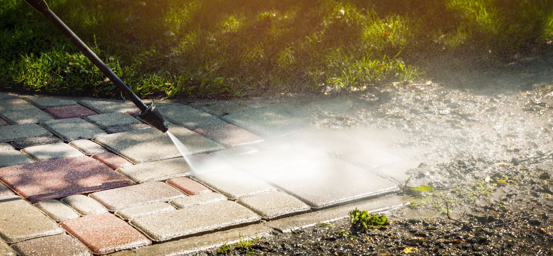 Person pressure-washing a sunlit paved patio outdoors, spraying water over wet bricks and grass edge.