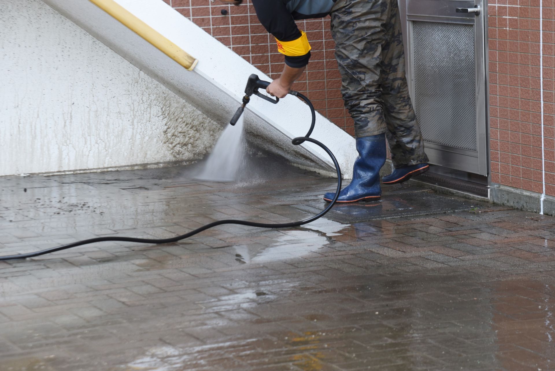 Person pressure-washing a wet sidewalk beside a building, wearing gloves and boots.