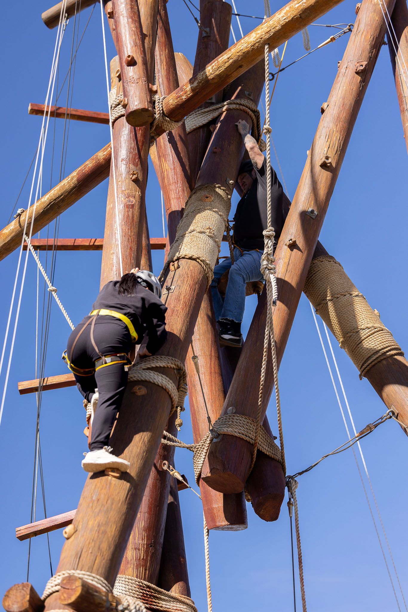 Two people climbing a wooden ropes course against a blue sky