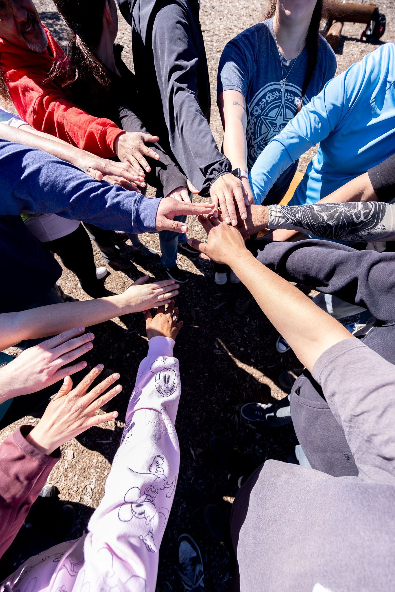 Hands of multiple people stacked together in a team huddle outdoors, with colorful sleeves visible.