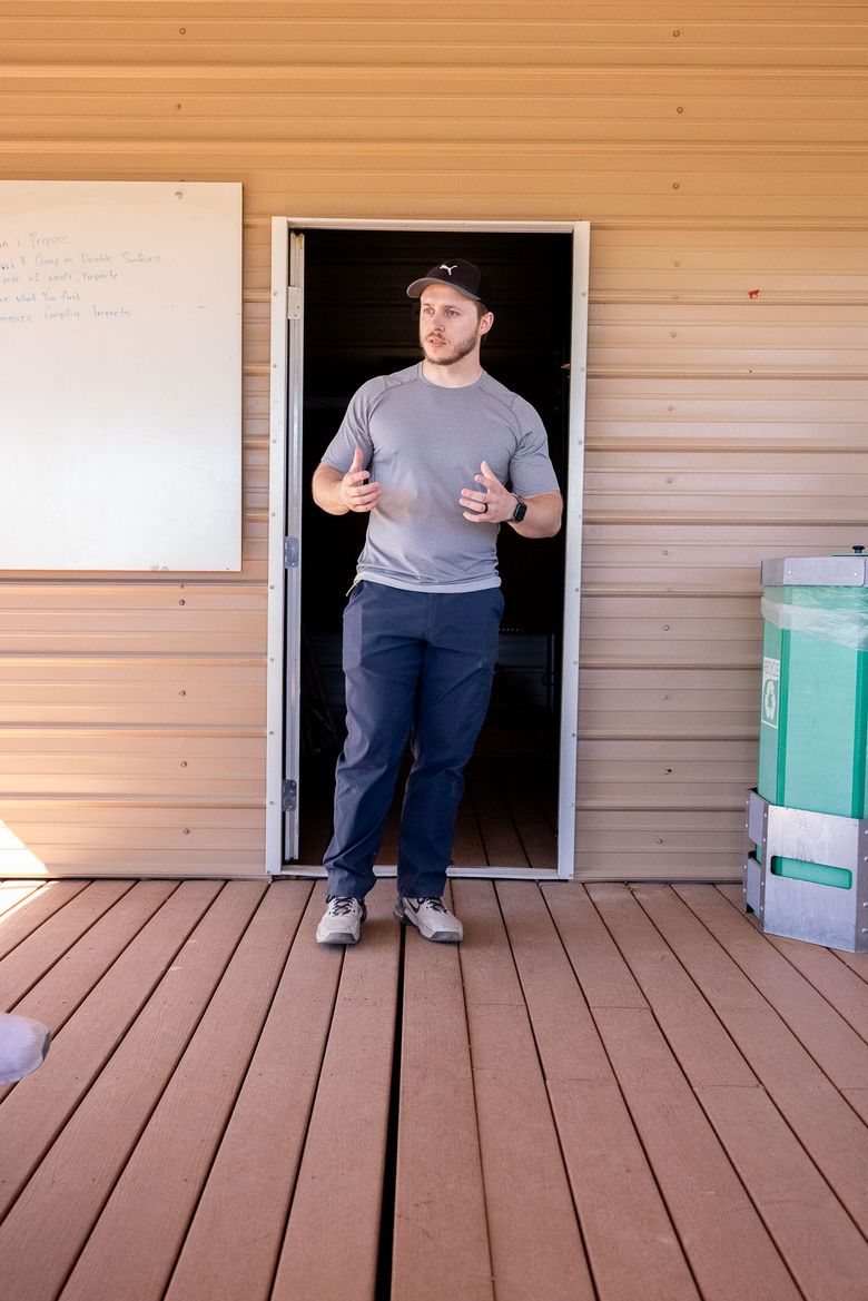 Person standing in a doorway on a wooden deck, gesturing while speaking, with a whiteboard nearby.