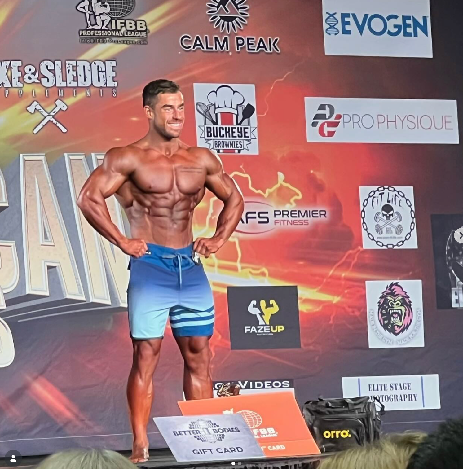 Bodybuilder poses on stage, wearing blue shorts. He is flexing, smiling, and surrounded by sponsor logos.