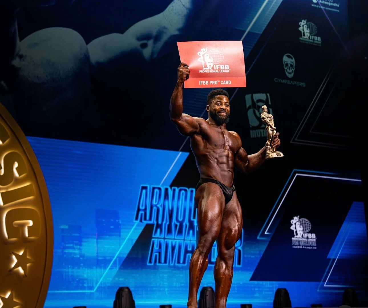 Female bodybuilder, stage, holding up a sign. She's wearing a teal bikini, muscles flexed, dark backdrop.