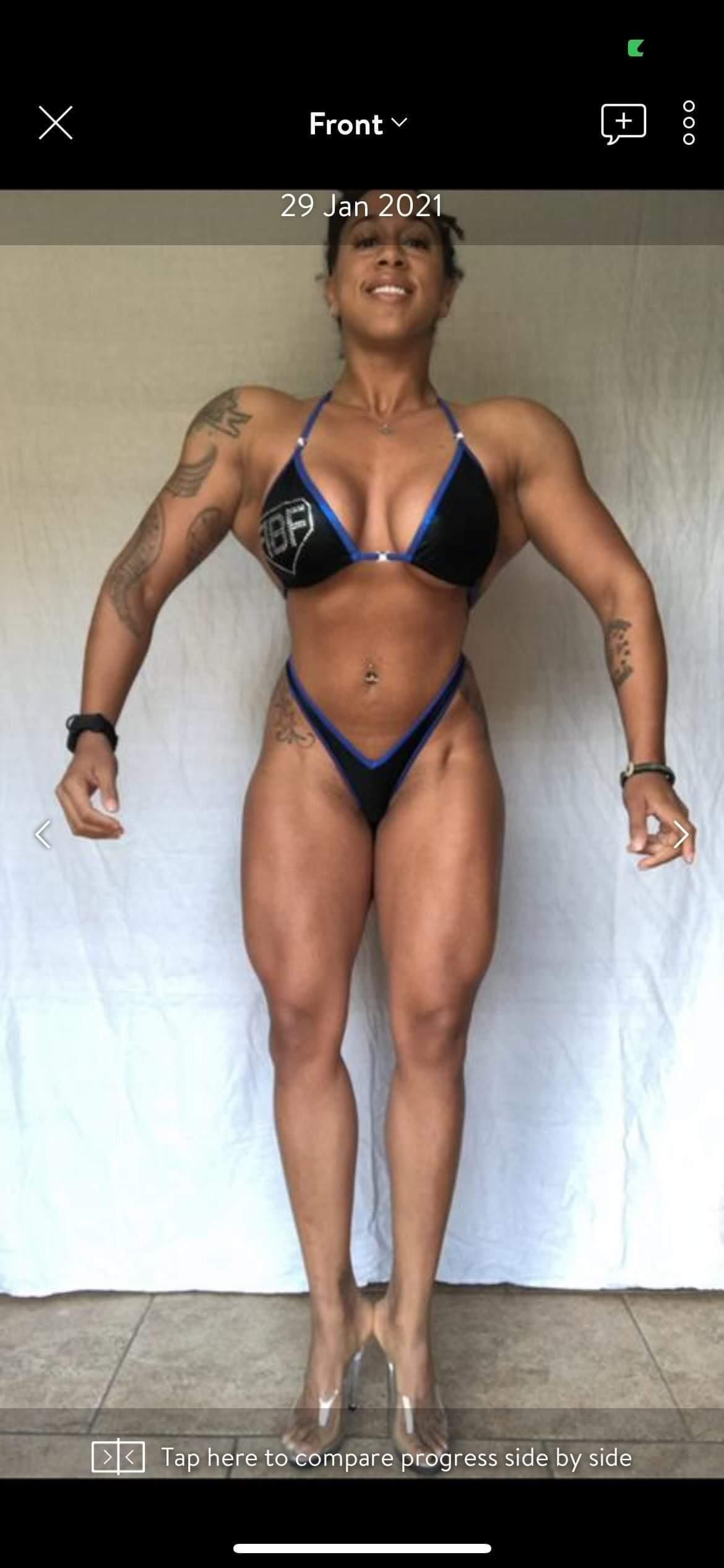 A bodybuilder in a bikini poses, showcasing muscular physique, arms extended, with a neutral backdrop.