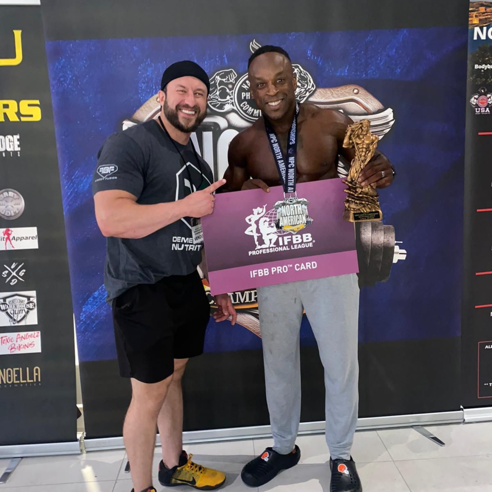 Two men, one holding a trophy and pro card, smiling, standing in front of a banner.