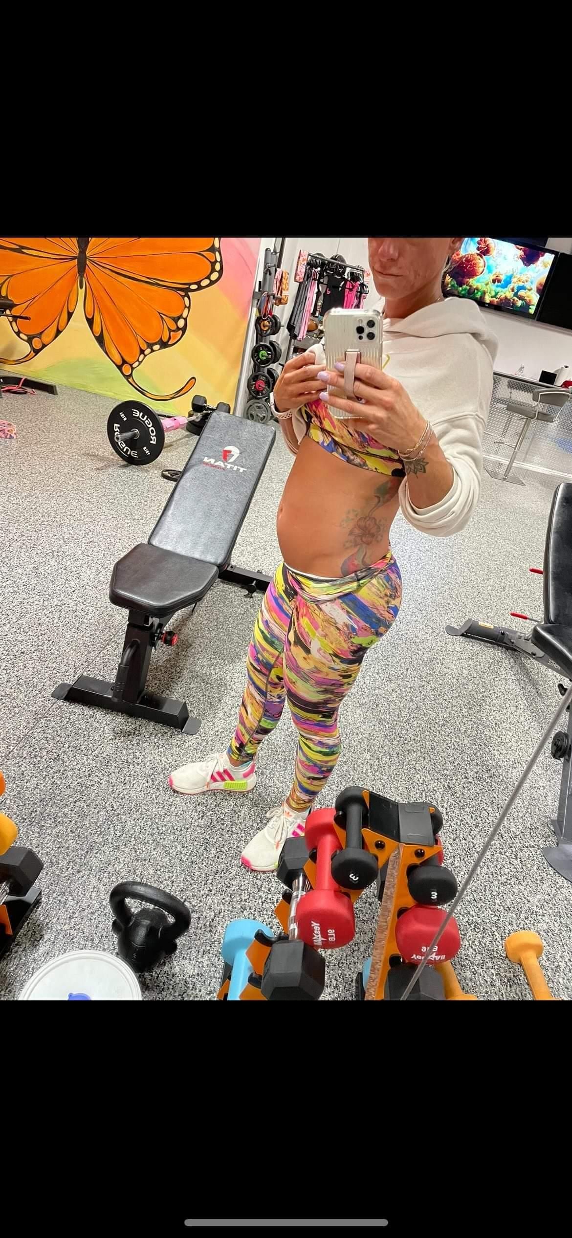 Woman in workout clothes takes a mirror selfie in a home gym, surrounded by weights and equipment.