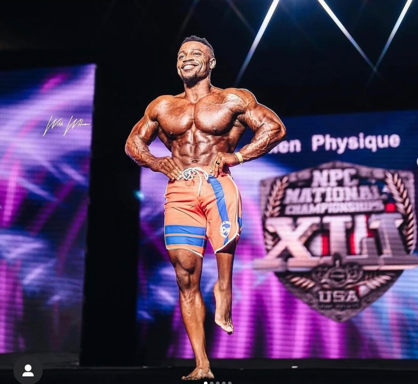 Bodybuilder on stage, posing in orange shorts. Shows defined muscles, smiling, at NPC Nationals.