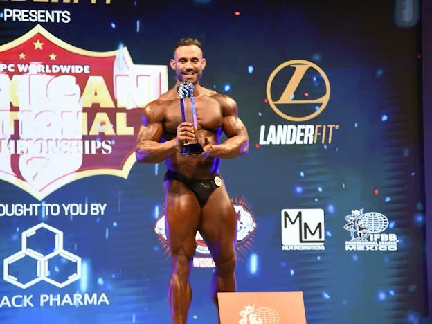 Bodybuilder on stage holding trophy, smiling. He's wearing posing trunks, in front of a banner at a fitness competition.